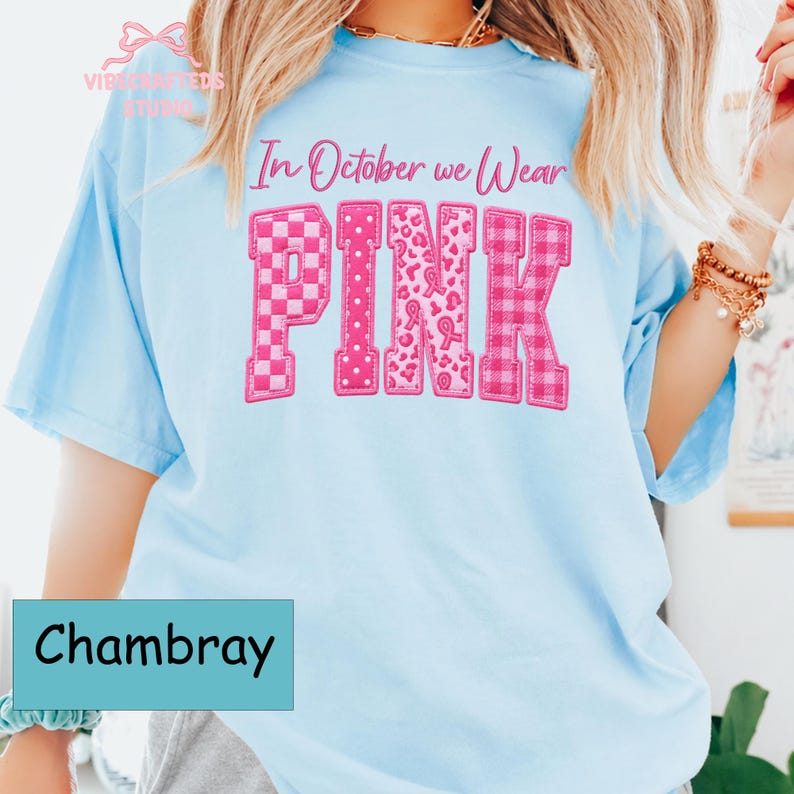 May include: A light blue short-sleeve t-shirt featuring the text In October we Wear PINK in pink lettering. The word PINK is displayed in large block letters with varied patterns including checkers, polka dots, leopard print, and plaid.