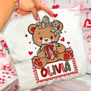 May include: White t-shirt featuring a cartoon teddy bear holding a heart-shaped gift. The bear has a bow and the name "OLIVIA" on a red and white sign. Red hearts and stars decorate the design. "VIBECRAFTED STUDIO" is printed on the top right.