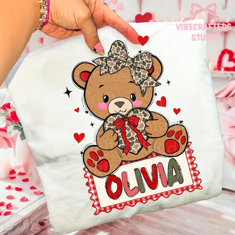 May include: White t-shirt featuring a cartoon teddy bear holding a heart-shaped gift. The bear has a bow and the name "OLIVIA" on a red and white sign. Red hearts and stars decorate the design. "VIBECRAFTED STUDIO" is printed on the top right.