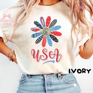 以下が含まれることがあります： 赤、青、黒の花びらを持つ大きな花の模様が特徴のアイボリー色のTシャツ。「USA」の文字が赤色の筆記体で花の下に書かれ、青い星がアクセントになっています。カジュアルなスタイルのシャツです。「IVORY」の文字は黒で書かれています。