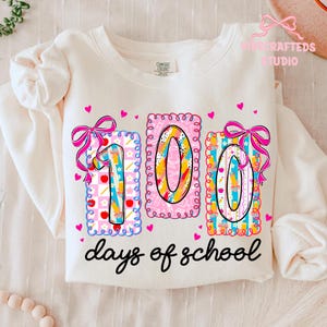May include: Cream-colored sweatshirt with a colorful "100 days of school" design. The design features the numbers "100" in decorative frames, bows, and hearts. The text "days of school" is written in a black script font.