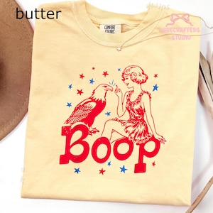May include: Cream-coloured t-shirt with a red and blue graphic. The graphic features an eagle, a woman, and the word "Boop" in large red letters. Blue and red stars are scattered around the design. The shirt has a crew neck and short sleeves.