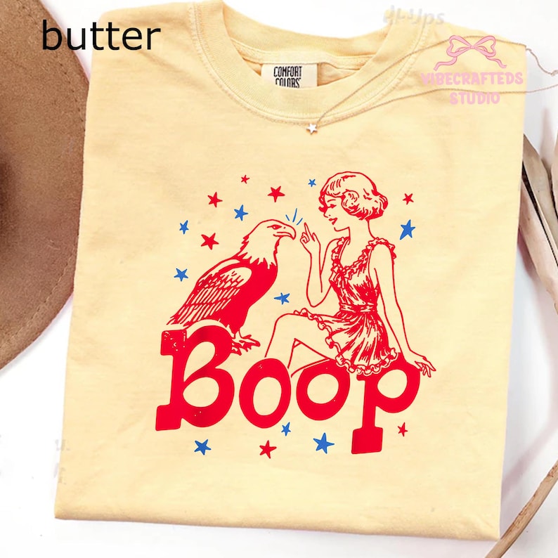 May include: Cream-coloured t-shirt with a red and blue graphic. The graphic features an eagle, a woman, and the word "Boop" in large red letters. Blue and red stars are scattered around the design. The shirt has a crew neck and short sleeves.
