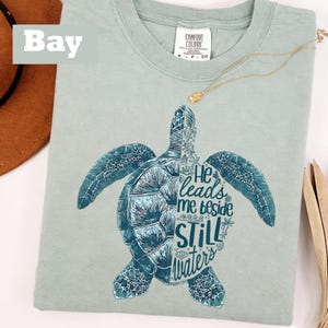 Op de afbeelding: Lichtgroen T-shirt met een blauwe zeeschildpad-afbeelding. Het schildpadontwerp bevat de tekst "He leads me beside still waters" en komt uit Psalm 23:2. Een gouden ketting is over het shirt gedrapeerd.