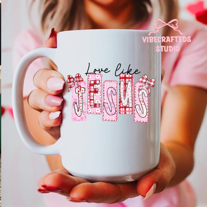 May include: A white ceramic mug with the words "Love like Jesus" printed on it. The word "Jesus" is in pink with decorative patterns and bows. The mug has a large handle and is held by a person.