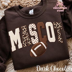 May include: A dark chocolate brown crewneck sweatshirt featuring the word MASCOT in large, textured chenille-style letters. The letters alternate between cream, brown, and leopard print patterns. A brown football graphic sits below the text.