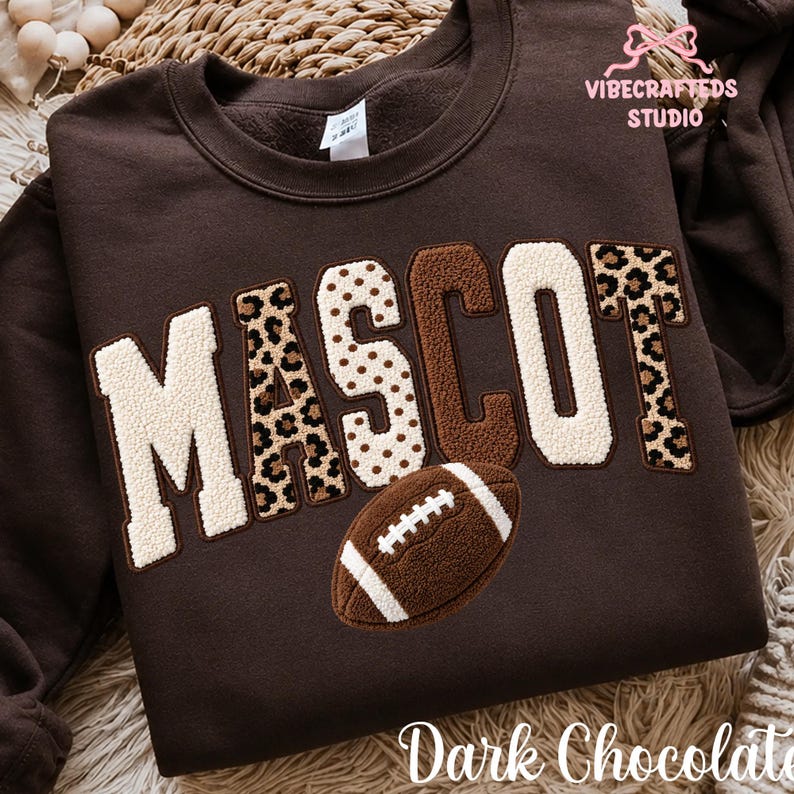 May include: A dark chocolate brown crewneck sweatshirt featuring the word MASCOT in large, textured chenille-style letters. The letters alternate between cream, brown, and leopard print patterns. A brown football graphic sits below the text.