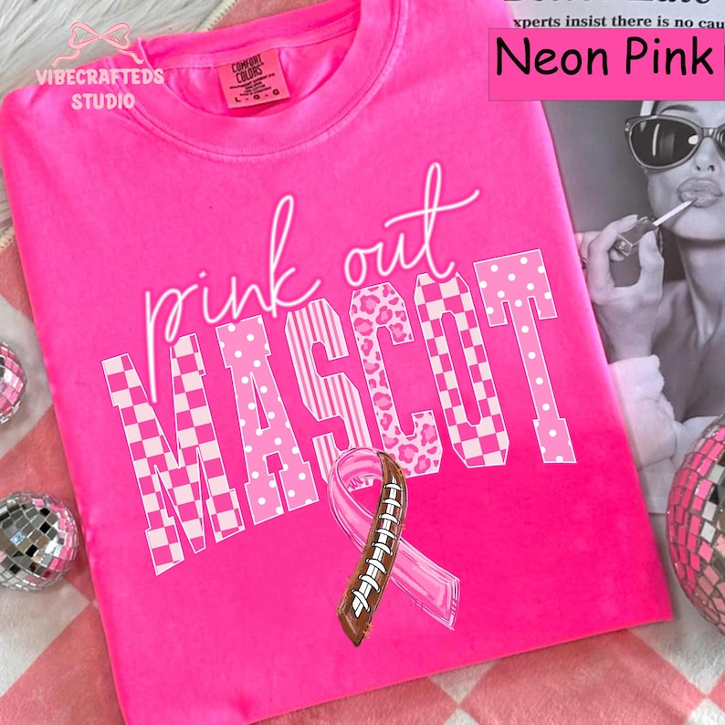 May include: A neon pink crew neck t-shirt featuring the white text "pink out MASCOT" in a mix of checkered, polka dot, and leopard print patterns. Below the text is a pink awareness ribbon with a brown football lace design in the center.