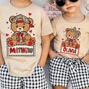 May include: Two tan t-shirts with teddy bear graphics. One shirt features a bear wearing a camo hat and holding a heart with the name "MATHEW" on a sign. The other shirt has a bear with a bow and the name "OLIVIA". Both shirts have red hearts.