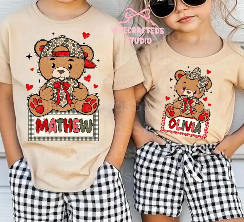 May include: Two tan t-shirts with teddy bear graphics. One shirt features a bear wearing a camo hat and holding a heart with the name "MATHEW" on a sign. The other shirt has a bear with a bow and the name "OLIVIA". Both shirts have red hearts.