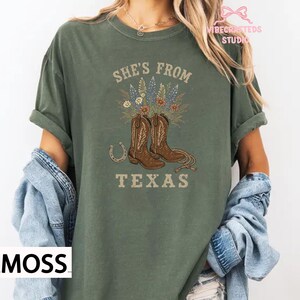 May include: Moss green t-shirt with the text "She's From Texas" and a graphic of cowboy boots, flowers, and a horseshoe. The shirt has short sleeves and a crew neck.