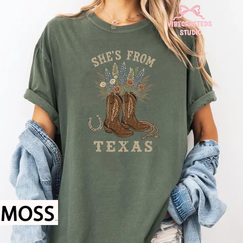 May include: Moss green t-shirt with the text "She's From Texas" and a graphic of cowboy boots, flowers, and a horseshoe. The shirt has short sleeves and a crew neck.