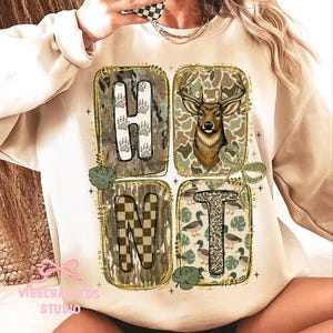 Pode incluir: Camisola creme com um design gráfico com a palavra "HUNT" num layout quadrado. As letras são preenchidas com padrões de camuflagem, pegadas, um veado e patos. Glitter dourado contorna os quadrados.