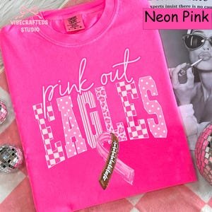 May include: A neon pink t-shirt featuring the text "pink out EAGLES" in white with checkered and leopard print patterns. A pink awareness ribbon with a brown football lace design is centered below the text. The shirt has a crew neckline.