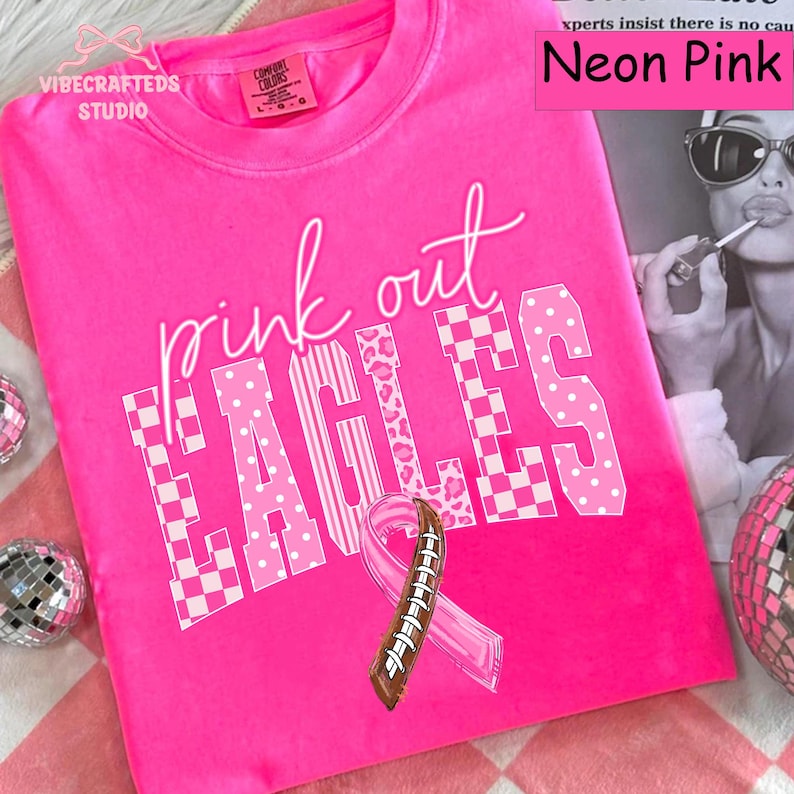 May include: A neon pink t-shirt featuring the text "pink out EAGLES" in white with checkered and leopard print patterns. A pink awareness ribbon with a brown football lace design is centered below the text. The shirt has a crew neckline.