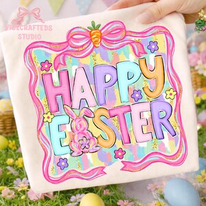 May include: A cream-colored tote bag with a colorful "Happy Easter" design. The design features pastel letters, a pink bunny, and floral accents within a pink and gold frame. A pink bow with a carrot is at the top.
