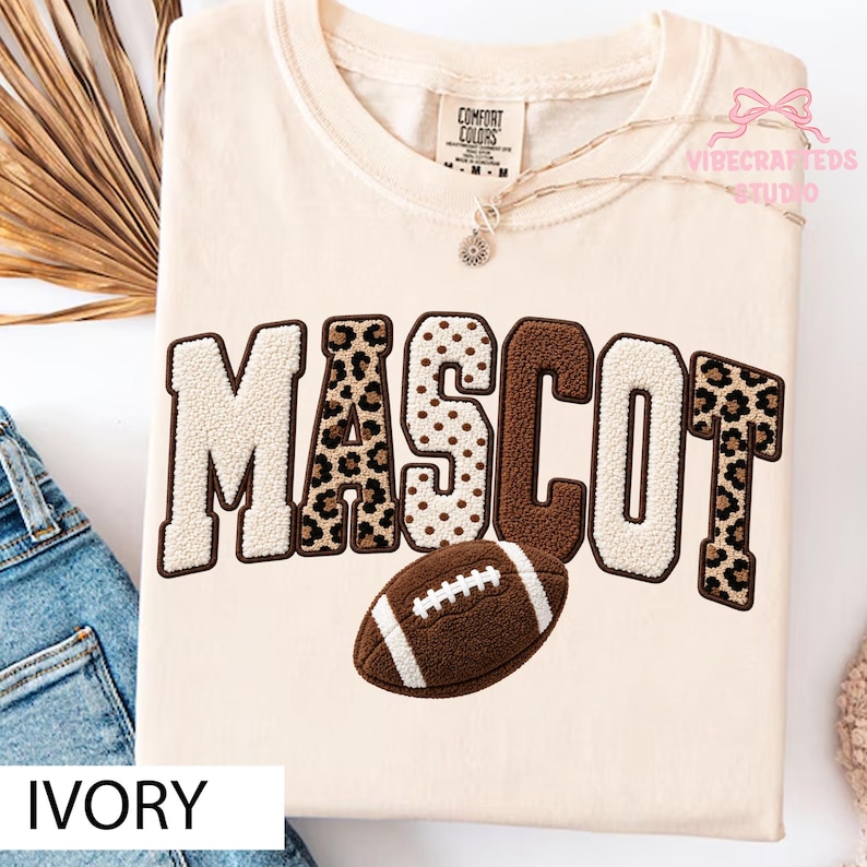 May include: An ivory t-shirt featuring the word MASCOT in large, textured, chenille-style letters with varying patterns including leopard print, polka dots, and solid brown. A brown football graphic is positioned below the text.