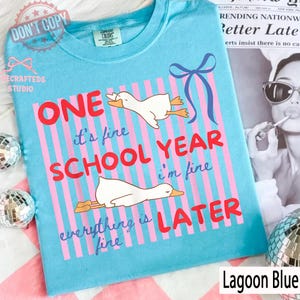 May include: Lagoon blue t-shirt with a whimsical design. The shirt features pink vertical stripes, cartoon geese, and the text "ONE SCHOOL YEAR LATER." A blue bow graphic is also present. The shirt is labeled "Lagoon Blue."