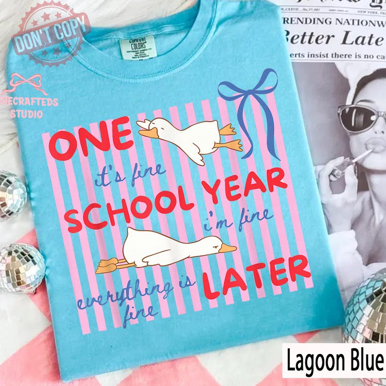 May include: Lagoon blue t-shirt with a whimsical design. The shirt features pink vertical stripes, cartoon geese, and the text "ONE SCHOOL YEAR LATER." A blue bow graphic is also present. The shirt is labeled "Lagoon Blue."