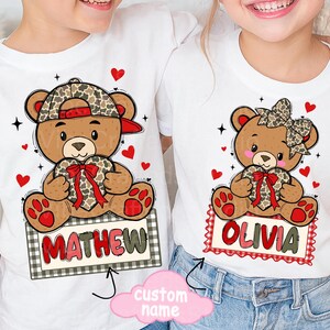 May include: White t-shirts featuring cartoon teddy bears holding heart-shaped gifts. One bear wears a camo hat and the name "MATHEW" on a sign. The other bear has a bow and the name "OLIVIA". Red hearts and stars decorate the shirts.