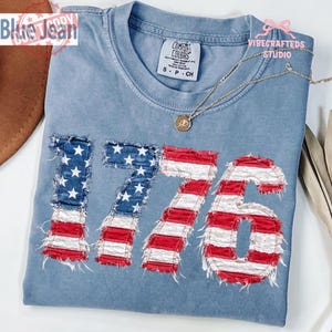 May include: A blue-grey t-shirt with the number "1776" appliqued on the front in a distressed American flag design. The shirt has a crew neck and short sleeves. A gold necklace with a pendant rests on the shirt.