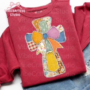 May include: A maroon sweatshirt with a colorful patchwork cross design. The cross features a variety of patterned fabrics, including floral, plaid, and solid colors, with a large bow at the center. The brand name "VIBECRAFTED STUDIO" is printed on the top left.