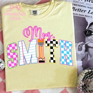 May include: A light yellow t-shirt with the name "Mrs. SMITH" in a colorful, playful font. The letters have checkerboard and pencil-themed designs. The shirt has the "Comfort Colors" brand tag.