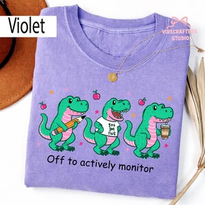 May include: A violet-colored t-shirt featuring three cartoon dinosaurs. The dinosaurs are green with pink accents and are holding a pencil, wearing a shirt that says "You got this," and holding a coffee cup. The text "Off to actively monitor" is printed at the bottom.