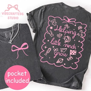 May include: Two charcoal gray t-shirts. One has a pink bow graphic, the other has the phrase "Helping little minds grow" in pink, surrounded by doodles. The text "pocket included" is in a pink circle. A silver chain necklace is on the first shirt.
