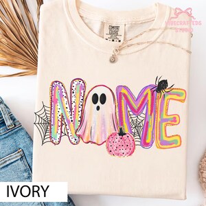 May include: An ivory t-shirt featuring a colorful graphic with the word NAME, where the letter A is replaced by a pastel ghost. The design includes a pink pumpkin, black spiderwebs, and a small black spider, all printed on a cotton fabric.