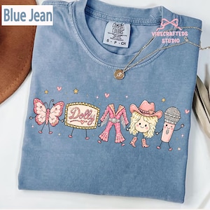 May include: A blue jean colored t-shirt featuring a graphic print of a butterfly, a marquee sign with the text Dolly, a pink fringe outfit, a blonde character wearing a cowboy hat, and a microphone, all holding hands with small stars and hearts.