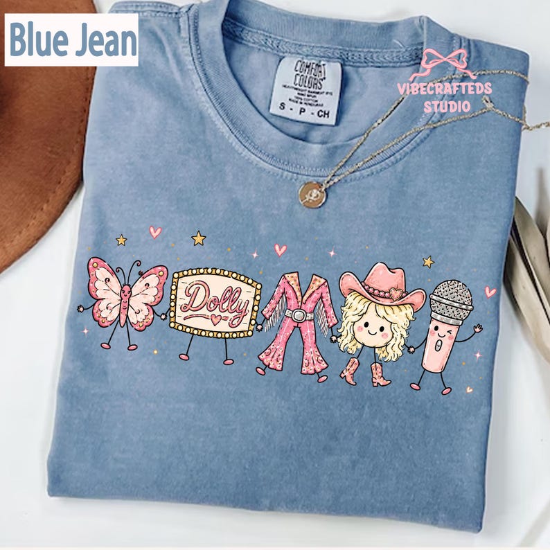 May include: A blue jean colored t-shirt featuring a graphic print of a butterfly, a marquee sign with the text Dolly, a pink fringe outfit, a blonde character wearing a cowboy hat, and a microphone, all holding hands with small stars and hearts.