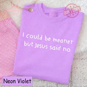 May include: A neon violet t-shirt with the text "I could be meaner but jesus said no" in white. The shirt has a crew neck and is folded. A pink gingham fabric is visible on the left.