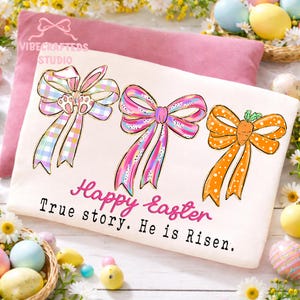 Puede incluir: Una camiseta crema con temática de Pascua con tres lazos decorativos. Los lazos son de colores pastel, uno diseñado como orejas de conejo y otro como zanahoria. El texto "Happy Easter" y "True story. He is Risen." está escrito en rosa y negro.