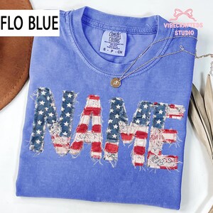 May include: A blue t-shirt with the word "NAME" appliqued in a patriotic American flag design. The shirt has a crew neck and short sleeves. The text "FLO BLUE" is in the upper left corner. A silver necklace with a pendant is on the shirt.
