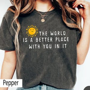 May include: A charcoal gray short-sleeve t-shirt featuring a yellow sun graphic with a smiling face and the white text "THE WORLD IS A BETTER PLACE WITH YOU IN IT" printed on the front. The fabric has a washed, vintage-style texture.