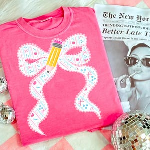 May include: A bright pink t-shirt featuring a large bow design with a pencil graphic. The bow is decorated with a pattern of apples, ribbons, and other school-themed elements. The text on the newspaper reads "TRENDING NATIONWIDE Better Late T."