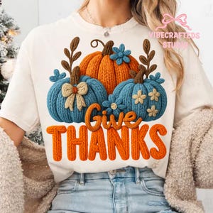 May include: Cream-colored t-shirt featuring a Thanksgiving design. The design includes three knitted pumpkins in orange and blue, with the words "Give Thanks" in orange. The shirt has a crew neck and short sleeves.