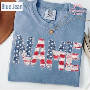 May include: A light blue t-shirt with the word "NAME" appliqued in a patriotic design. The letters feature a combination of red and white stripes, denim with white stars, and lace. The shirt has a crew neck and short sleeves.