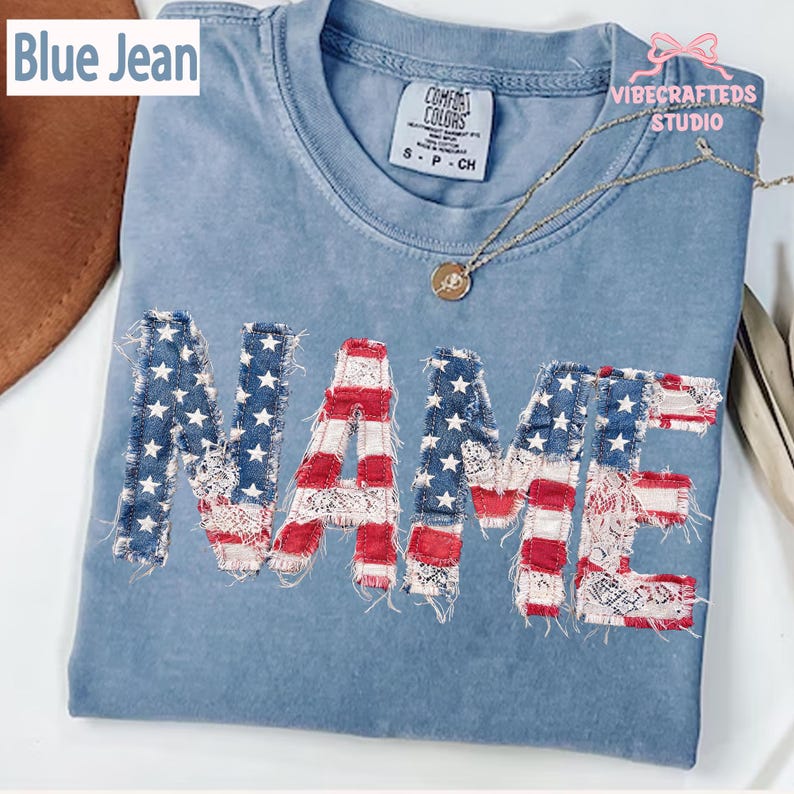 May include: A pale blue t-shirt with the word "NAME" appliqu&eacute;d in a patriotic design. The letters feature a combination of red and white stripes, denim with white stars, and lace. The shirt has a crew neck and short sleeves.