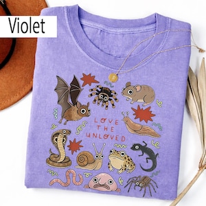 May include: A violet t-shirt with illustrations of various creatures, such as a bat, spider, and snake, and the text "LOVE THE UNLOVED". A gold necklace and a brown hat are also visible in the image.