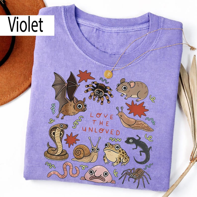 May include: A violet t-shirt with illustrations of various creatures, such as a bat, spider, and snake, and the text "LOVE THE UNLOVED". A gold necklace and a brown hat are also visible in the image.