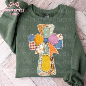 May include: Olive green sweatshirt with a patchwork cross design. The cross features a colorful bow and various fabric patterns, including plaid and floral prints. The text "VibeCrafteds Studio" is visible.