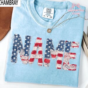 May include: Pale blue t-shirt with the word "NAME" appliqu&eacute;d in a patriotic design. The letters feature red and white stripes, blue with white stars, and lace details. A silver necklace with a pendant rests on the shirt.