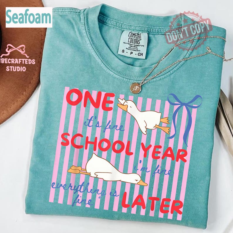 May include: Seafoam green t-shirt with the text "ONE SCHOOL YEAR LATER" in red and pink. The shirt features cartoon ducks and a blue bow design. The shirt is a Comfort Colors brand, size S.