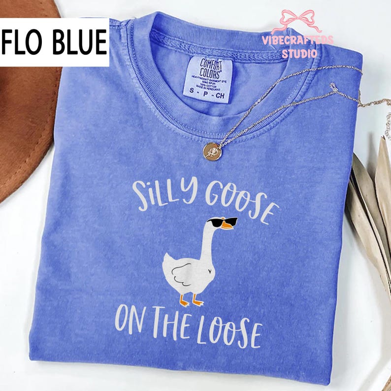 May include: A Flo Blue Comfort Colors t-shirt with the text "Silly Goose On The Loose" and a graphic of a goose wearing sunglasses. A gold necklace with a pendant is draped across the shirt.