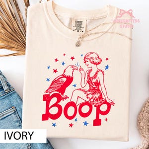 May include: Ivory Comfort Colors t-shirt featuring a red graphic of a woman, an eagle, and the word "Boop." The design includes red and blue stars. A silver necklace is visible. The shirt is displayed on a flat surface.