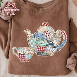 May include: A brown sweatshirt featuring a patchwork design of a teapot and teacup. The teapot and teacup are adorned with diverse fabrics in blue, red, and white patterns. The text "VIBECRAFTED STUDIO" is visible on the upper left.