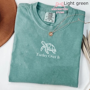 May include: Light green t-shirt with a white turtle graphic and the text "Turley Over It." The shirt has a crew neck and a small tag on the collar. A silver necklace with a round pendant is on the shirt.