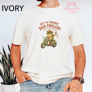 Puede incluir: Camiseta de color marfil con el texto "OFF TO COMMIT DAD FOOLERY" y una ilustración de una rana en un scooter con un lazo. La rana lleva sombrero de vaquero y botas. La camiseta tiene mangas cortas.